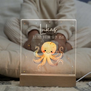 Peut inclure: Une veilleuse en acrylique transparente avec une base en bois. La lumière présente une illustration aquarelle d'une pieuvre avec un visage mignon. Le texte "Lukas 13.05.2023 - 18:43 Uhr - 50 cm - 3850g" est imprimé sur la lumière.
