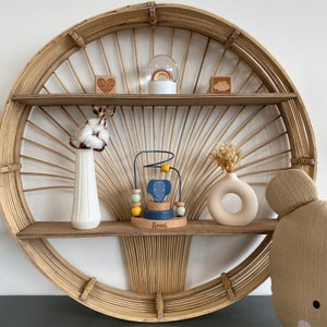 May include: A wooden shelf with a circular rattan frame, featuring a blue and yellow elephant toy, a white vase with cotton balls, a white vase with dried flowers, a wooden heart, a snow globe, and a wooden cloud.