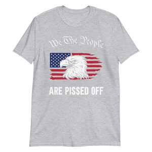 Pode incluir: Uma camiseta cinza com um design de bandeira americana desbotada e uma &aacute;guia. O texto "We The People Are Pissed Off" &eacute; impresso em branco.