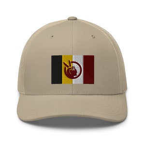 Puede incluir: Una gorra de camionero beige con una bandera bordada en negro, amarillo, blanco y rojo que presenta una mano estilizada y un círculo. La bandera está centrada en la parte delantera de la gorra.