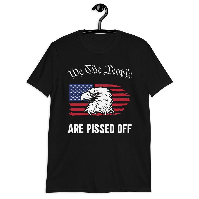 Pode incluir: Camiseta preta com um design de bandeira americana desbotada com uma &aacute;guia careca e o texto "We The People Are Pissed Off."