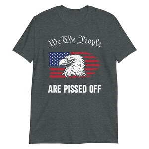 Pode incluir: Uma camiseta cinza escuro mesclada com um design de bandeira americana desbotada e uma cabe&ccedil;a de &aacute;guia. O texto "We The People Are Pissed Off" &eacute; impresso em branco.
