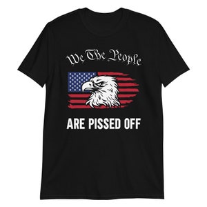 Pode incluir: Camiseta preta com um design de bandeira americana desbotada e uma cabe&ccedil;a de &aacute;guia. O texto "We The People Are Pissed Off" &eacute; impresso em branco abaixo da bandeira.