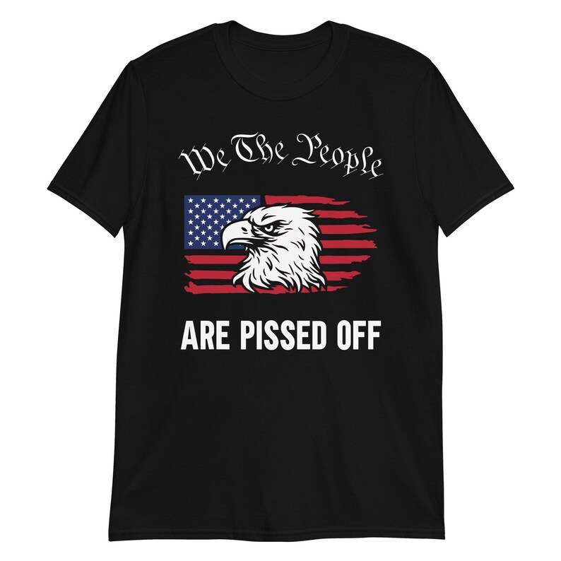 Pode incluir: Camiseta preta com um design de bandeira americana desbotada e uma cabe&ccedil;a de &aacute;guia. O texto "We The People Are Pissed Off" &eacute; impresso em branco abaixo da bandeira.
