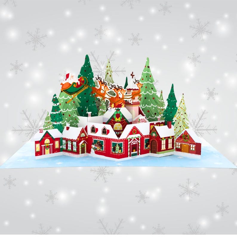May include: A 3D pop-up Christmas card featuring a winter village scene with Santa Claus flying over a snowy town with reindeer. The card is decorated with red and green houses, trees, and snow.