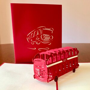 May include: A red 3D pop-up card featuring a red double-decker bus. The card is closed and shows a silhouette of a bus on the front.
