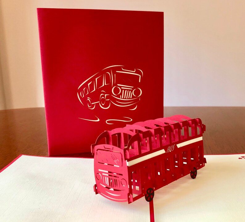 May include: A red 3D pop-up card featuring a red double-decker bus. The card is closed and shows a silhouette of a bus on the front.