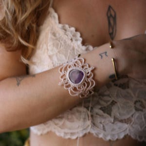 Könnte beinhalten: Ein zartes gehäkeltes Armband mit einem violetten Edelstein in der Mitte. Das Armband ist aus weißem Garn gefertigt und hat ein florales Design.