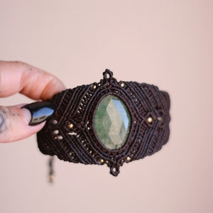 Puede incluir: Un brazalete de macramé marrón con una piedra preciosa verde en el centro. El brazalete está decorado con pequeñas cuentas doradas.