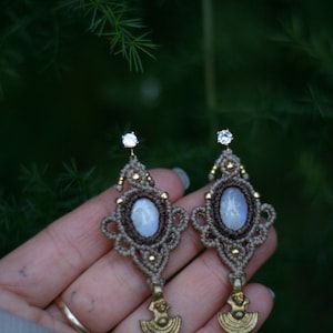 May include: A pair of handmade earrings featuring a moonstone gemstone set in a brown and gold macrame design. The earrings have a crescent moon charm hanging from the bottom.