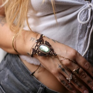Pode incluir: Uma pulseira de mão em macramé castanho com uma pedra preciosa de labradorite oval central com reflexos iridescentes verdes e azuis. A peça inclui um trabalho intrincado de cordão atado, pequenas contas metálicas e uma delicada corrente dourada.
