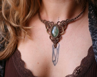 Puede incluir: Un collar de macramé hecho a mano con un gran colgante ovalado de piedra verde. El intrincado diseño de macramé marrón incluye detalles dorados y un dije colgante en forma de cristal. El collar se lleva sobre un top marrón.