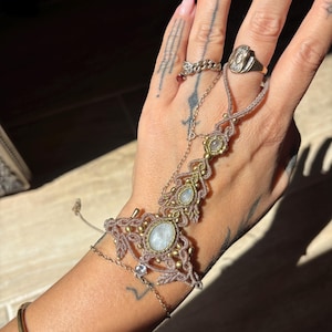 Puede incluir: Una cadena de mano con un diseño de cordón de macramé color topo, tres cabujones de piedra lunar iridiscentes de forma ovalada y pequeñas cuentas doradas. La pieza conecta una pulsera con un anillo, acentuada por una fina cadena dorada.