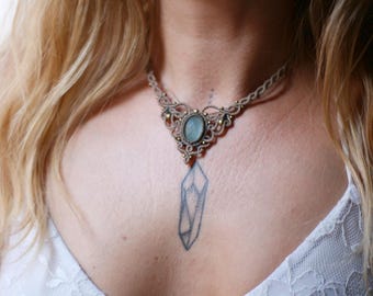 May include: A macrame necklace featuring a central oval light blue stone set in beige cord with small metallic beads. The intricate knotted design forms a decorative collar around the neck, showcasing detailed craftsmanship and a neutral color palette.