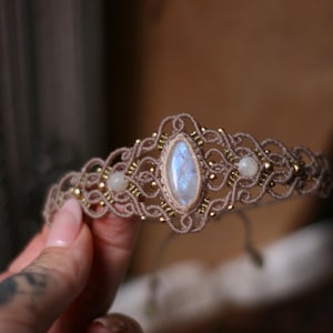 May include: A beige macrame bracelet featuring a central marquise-shaped iridescent moonstone cabochon, flanked by two smaller round moonstone beads and accented with small metallic gold-toned beads woven into the intricate knotted cord pattern.