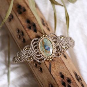May include: A macrame bracelet featuring a central oval cabochon with a blue and green marbled pattern. The beige cord is intricately knotted in a symmetrical pattern and accented with small metallic gold beads along the edges of the design.