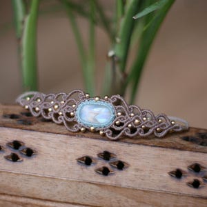 Peut inclure: Un bracelet en macramé avec une pierre de lune ovale irisée au centre, sertie dans un fil bleu clair. Le bracelet est composé d'un cordon couleur taupe avec des motifs de nœuds complexes et de petites perles dorées métalliques intégrées.
