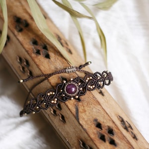 Op de afbeelding: Een donkerbruine macramé armband met een centrale ronde paarse edelsteen omringd door kleine metalen gouden kralen. Het ingewikkelde geknoopte koordontwerp bevat een verstelbare schuifsluiting met dunne bruine koordjes en gouden eindkralen.