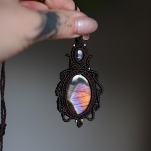 May include: A brown macrame necklace with a large iridescent gemstone pendant. The pendant is oval shaped and has a dark brown, purple, and orange color.