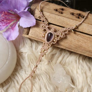Pode incluir: Uma joia de pé de macramé bege com contas douradas e uma pedra de ametista oval no centro. A joia é exibida em uma superfície de madeira com um fundo macio e branco fofo e uma flor roxa.