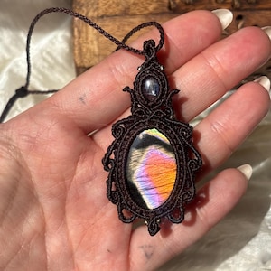 May include: A macrame pendant necklace featuring a large oval labradorite gemstone with iridescent orange, purple, and yellow flashes. The stone is encased in dark brown knotted cord with intricate decorative loops and a smaller dark stone accent.