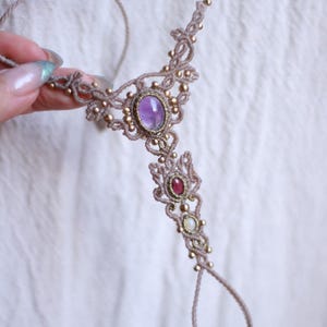 May include: A beige macrame hand chain featuring a central oval purple amethyst cabochon, a smaller red stone, and a tiny white stone. The intricate knotted cord design is accented with small metallic gold beads and includes a finger loop.