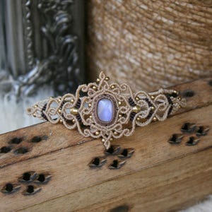 May include: A macrame bracelet featuring a central oval-shaped iridescent moonstone cabochon, surrounded by intricate beige and dark brown knotted cord patterns, accented with small metallic gold beads throughout the woven design.