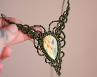 Puede incluir: Un collar de macramé hecho a mano con una piedra de labradorita en forma de lágrima. El collar está hecho con cordón verde oliva y adornado con pequeñas cuentas doradas. El intrincado trabajo de nudos rodea la piedra iridiscente.