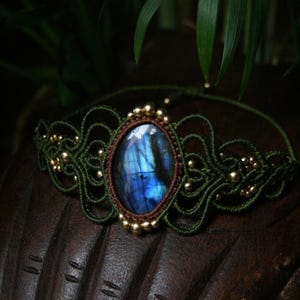 Peut inclure: Un collier ras du cou en macramé fait à la main, orné d'une grande pierre de labradorite ovale aux reflets bleus et verts. La pierre est sertie dans un cadre marron, entourée de perles dorées et de détails en macramé vert olive.