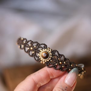 Könnte beinhalten: Ein dunkelbraunes Makramee-Armband mit einem zentralen, runden Tigerauge-Edelstein in einer goldfarbenen Metallfassung. Das geflochtene Kordeldesign ist mit kleinen, gleichmäßig verteilten goldfarbenen Perlen verziert, die in das Knotenmuster eingearbeitet sind.