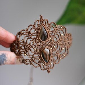 May include: A brown macrame bracelet with two tiger's eye stones. The bracelet is intricately woven with a delicate pattern and features gold beads.