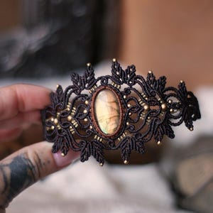Könnte beinhalten: Ein dunkelbraunes Makramee-Armband mit einem ovalen, irisierenden Labradorit-Edelstein in der Mitte. Das komplexe Design aus geknoteten Schnüren ist mit kleinen, metallisch goldenen Perlen verziert, die symmetrisch im Muster angeordnet sind.