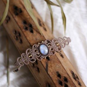 Puede incluir: Una pulsera de macramé con una piedra lunar ovalada central que presenta un brillo iridiscente azul. La pulsera está hecha de cordón marrón claro con un patrón de nudos intrincado, acentuado con pequeñas cuentas doradas metálicas.