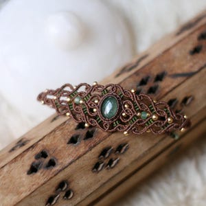 Puede incluir: Un pasador de pelo de macramé con cordón marrón y verde tejido en un patrón intrincado. El diseño está centrado alrededor de una piedra preciosa ovalada verde y acentuado con pequeñas cuentas doradas en toda la estructura.