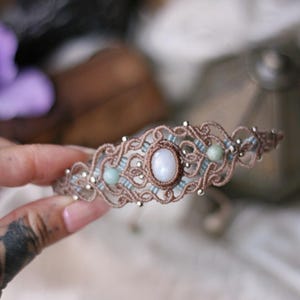 May include: A macrame bracelet featuring a central oval white stone, flanked by two smaller round light green beads. The design is crafted from brown cord with intricate knotted patterns, accented by small metallic silver beads throughout the piece.