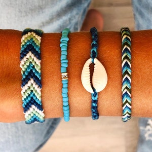 Peut inclure: Quatre bracelets aux tons de bleu, vert et blanc. Deux sont tissés avec des motifs en chevrons, l'un est une chaîne de perles turquoise, et le dernier présente un coquillage blanc. Ces bracelets faits à la main sont parfaits pour un style bohème.