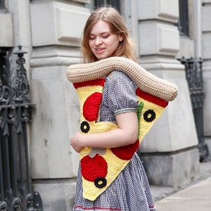 Puede incluir: Una mujer con un vestido de cuadros vichy lleva un gran bolso de pizza en forma de rebanada de crochet. El bolso es amarillo, rojo, verde y marrón con detalles negros.