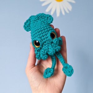 May include: A teal blue crocheted squid with large black eyes. The squid is being held in a hand against a light blue background.