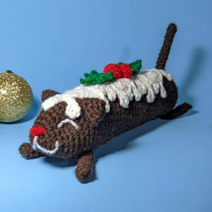 May include: A handmade crochet Christmas pudding cat with a brown body, white icing, and red berry and green leaf accents. The cat has a red nose and a white face. A gold glitter ornament is in the background.