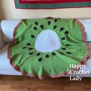 May include: A green and brown crocheted throw blanket shaped like a kiwi fruit. The blanket has a brown fringe around the edges and black crocheted seeds on the kiwi.