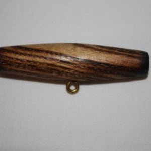 Könnte beinhalten: Ein Holz-Toggle mit einem dunkelbraunen und hellbraunen Streifenmuster. Der Toggle hat eine kleine Messingöse an der Unterseite.