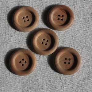 Peut inclure: Cinq boutons en bois avec quatre trous chacun. Les boutons sont de couleur brun clair et ont une surface lisse et arrondie.