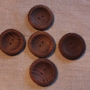 Peut inclure: Cinq gros boutons ronds en bois avec une finition marron foncé. Chaque bouton a deux trous et une surface texturée et striée. Les boutons sont disposés sur un fond en tissu de ton neutre.
