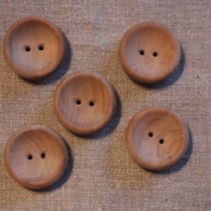 Puede incluir: Cinco botones redondos de madera, cada uno con dos agujeros, dispuestos sobre una tela beige texturizada. Los botones tienen una veta de madera natural y un diseño ligeramente cóncavo.