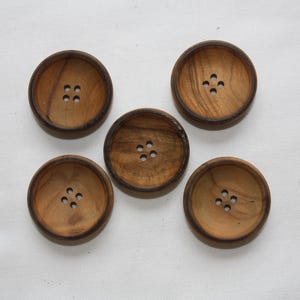 Peut inclure: Cinq boutons en bois avec une finition brun foncé et plusieurs trous. Les boutons sont ronds et ont un bord légèrement surélevé.
