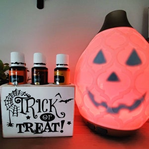 Peut inclure: Une boîte en bois blanche avec le texte "Trick or Treat!" et un motif de toile d'araignée. Quatre petites bouteilles en verre contenant un liquide brun sont dans la boîte. Un diffuseur en forme de citrouille avec une lueur rouge est en arrière-plan.