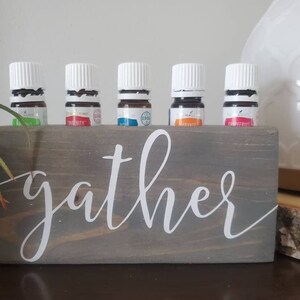 May include: A wooden essential oil diffuser holder with the word "gather" written in white script. The holder has six slots for essential oil bottles, and five bottles are in the slots. The bottles are all clear glass with white lids and contain different coloured liquids.