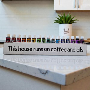 May include: A white wooden sign with the text "This house runs on coffee and oils" holds a row of small essential oil bottles. The bottles are in various colors, and a potted plant is in the background.