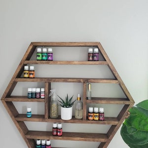 May include: A hexagonal wooden shelf, stained brown, with several compartments holding essential oil bottles, a spray bottle, a small potted plant, and a glass bottle with a dropper. The shelf is mounted on a light grey wall.