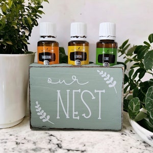 May include: A wooden sign with the text "our NEST" painted in white on a light green background. Three small glass bottles with white lids are displayed in a holder above the sign. The bottles are labeled "Tangerine", "Lemon", and "Lime".